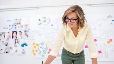 Žena u žutoj košulji sa naočarima sedi za stolom i piše. Iza nje se nalazi zid sa fotografijama, beleškama i skicama.