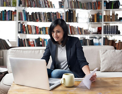 Woman sitting on a sofa, working on a laptop with documents in hand, a coffee mug on the table
