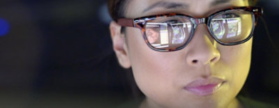 A focused person with glasses looks at a computer screen, reflections of data visible in the lenses.