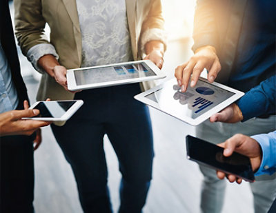 A group of people holding smartphones and tablets showing data charts and graphs, suggesting a collaborative business or tech discussion.