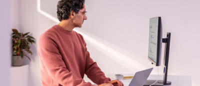 A man in a pink sweater works at a desk with a laptop and monitor.