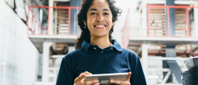 Smiling women in blue shirt holding a tablet in an industrial setting.
