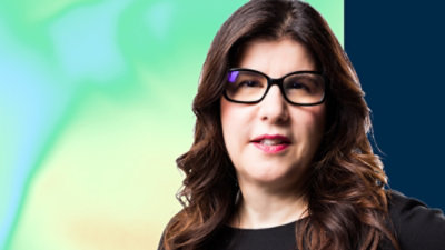 A person with long brown hair wearing glasses looking up at a green and blue sky.