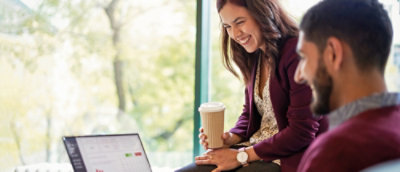 A woman is holding a cup and smiling, while a man who is also smiling is looking at a laptop.