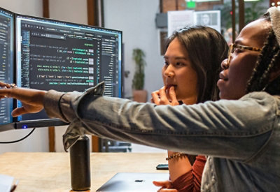 Two women engaged in a discussion while working on a computer with a monitor in view.