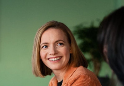 Person seated indoors wearing an orange sweater, turned slightly toward another person against a green background.