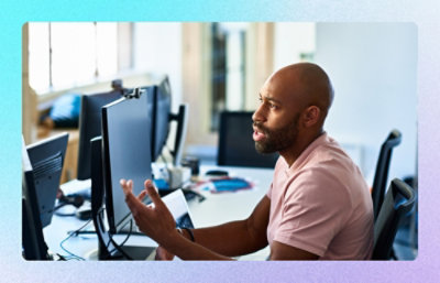 Person seated at desk discussing work while viewing multiple computer monitors in an office.