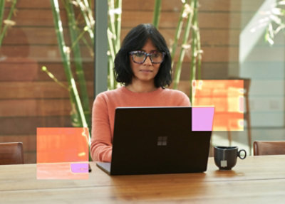 Una mujer con gafas y camisa rosa sentada en un escritorio con un portátil.