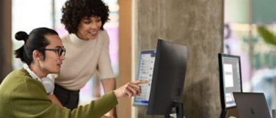 Una mujer con camisa blanca señala la pantalla de un ordenador mientras un hombre con gafas y jersey verde la observa.