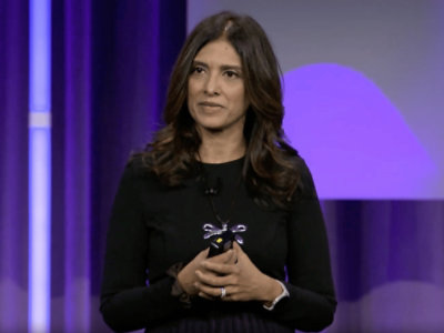 A woman speaking on stage holding clicker with purple backdrop.