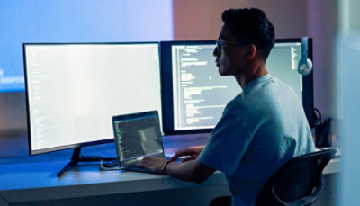 Person seated at a desk coding on a laptop with two large monitors displaying code.
