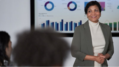 Eine lächelnde Frau mit ineinander verschränkten Händen, die vor einer Gruppe Personen steht.