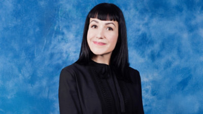 Person with straight, shoulder-length dark hair wearing a black blouse against a blue textured background
