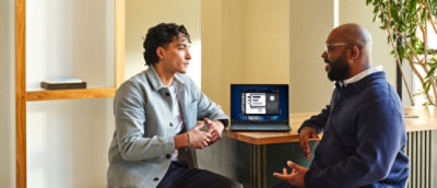 Two men sit at a table engaged in conversation. A laptop displaying software is open between them.