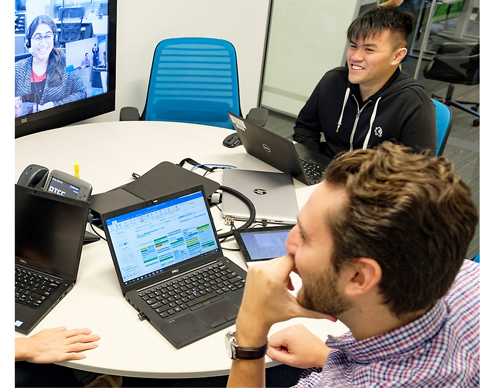 Dos hombres y una mujer en una videollamada participando en una animada reunión de negocios con ordenadores portátiles en una oficina moderna.