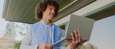 Person standing outdoors, holding and looking at an open laptop.