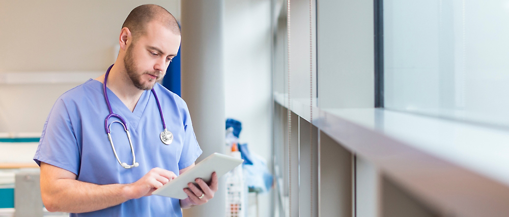 Male nurse in scrubs with a stethoscope reading a tablet in a hospital corridor.