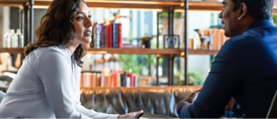Two people sitting and facing each other at a table in a room with bookshelves in the background.
