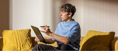 A person sitting on a couch using a tablet and stylus next to a yellow cushion.