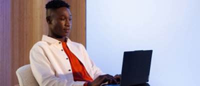  Person wearing a white button-up shirt over a bright orange top, seated and typing on a black laptop in a modern setting with light wooden panels and a plain backdrop.