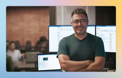 Person in a dark green T-shirt standing with arms crossed in front of multiple computer monitors displaying system dashboards in an office.