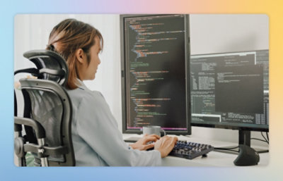 Person seated at a desk typing on a keyboard, with two large monitors displaying code in a modern workspace.