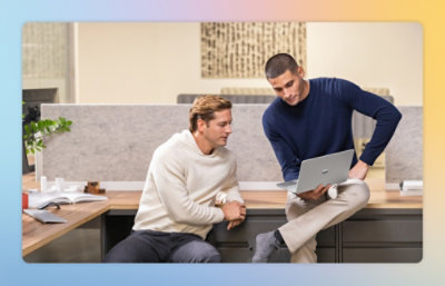Two people in a modern office sitting on a desk edge, one holding a laptop and discussing work with the other.