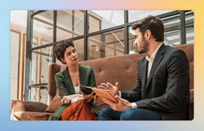 Two people sitting on a brown sofa in a modern office, holding tablets and engaged in a discussion.