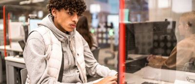 Person in a light vest and gray hoodie working at a computer in a modern office with red dividers.