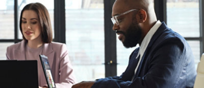  Two people sitting at a table with laptops in a modern office, engaged in work or discussion.