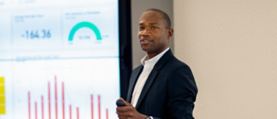 A person in a suit holding a microphone, standing in front of a screen with text -164 36.