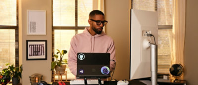 Person in a pink hoodie standing at a desk with a laptop and large monitor in a home office with framed art and plants.