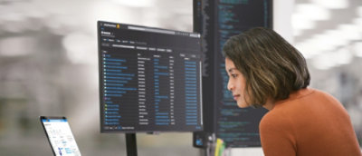 Person in an orange top working at a desk with multiple monitors displaying code and a laptop showing a calendar.