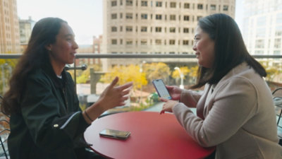 Two people sitting at a round red table outdoors, one holding a smartphone while the other gestures during a conversation, with city buildings in the background.