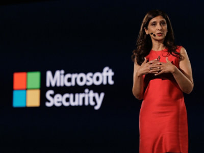 Presenter in a red outfit speaking on stage with Microsoft Security displayed on a screen in the background.
