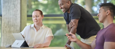 Three people collaborating around a tablet in a bright office setting. One is seated working; the other two are standing, discussing with a smile.