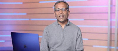 A man wearing glasses and a grey shirt stands in front of a blue wall with a logo.