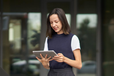 女性が屋外に立って、タブレット デバイスを持って見ています。彼女は、ノースリーブのトップスの下に白い T シャツを着ています。