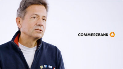 A man in a blue jacket stands in front of a building with the text COMMERZBANK.