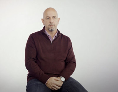 A man with a goatee wearing a purple shirt sitting on a chair.