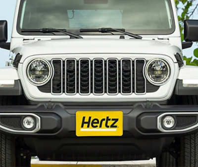 Front view of a white SUV with round headlights and grille, displaying a yellow Hertz license plate.