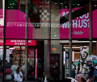 T‑Mobile storefront with pink branding and signage, viewed through glass with pedestrians in front.