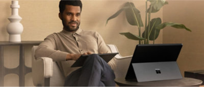 Een persoon in een beige trui zit op een bank met een tablet in de hand. Een geopende laptop staat op de tafel ernaast