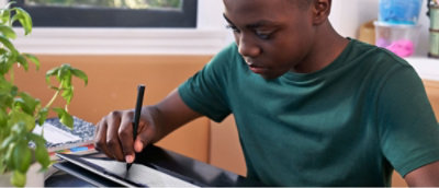 Person in a green shirt writing in a notebook at a desk with a potted plant nearby.