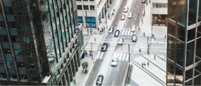 Aerial view of a city street with a few cars and pedestrians at a crosswalk, flanked by tall buildings.
