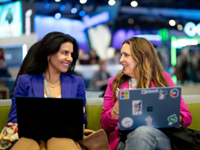 Two people seated at a technology event, working on laptops with exhibition booths in background