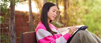 A woman sitting on a bench writing on a tablet.