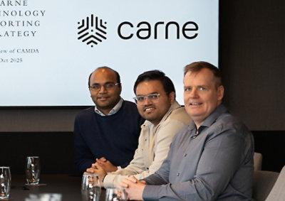 A group of men sitting at a table with visible text on a screen in the background.