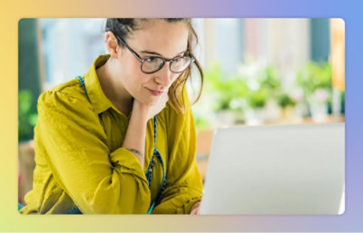 A woman wearing glasses and a yellow shirt is looking at a laptop on a white surface.