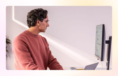 Person in a pink sweater sitting at a desk looking at a monitor with a laptop in front.